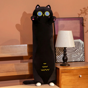 Op een bed met daarnaast een nachtkastje met een wit bedlampje en een frame erachter ligt een pluche zwart kattenkussen, het staat op zijn kleine achterpootjes