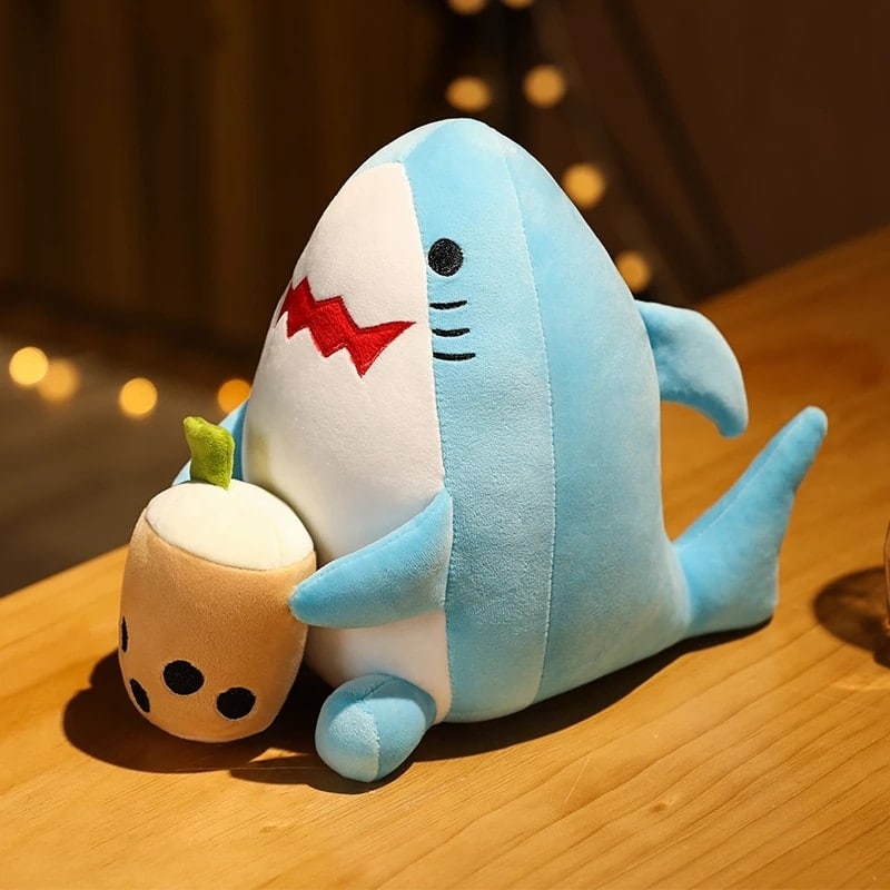 Bellentheepluche in de vorm van een blauwe haai zittend op een houten tafel met een kleine beige pot in zijn handen